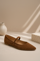 A single brown suede Mary Jane flat shoe with a pointed toe, displayed on a beige surface with a white ceramic vase and a textured stone block in the background.