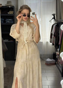 Woman in a beige dress taking a mirror selfie in a room with clothing and furniture.