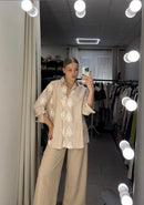 A woman takes a mirror selfie in a fitting room wearing a beige short-sleeve button-up shirt with white embroidered patterns and matching beige wide-leg trousers.