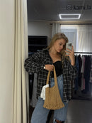 A woman takes a mirror selfie in a fitting room, wearing a black and white plaid flannel shirt, blue jeans, and holding a large woven straw shoulder bag.