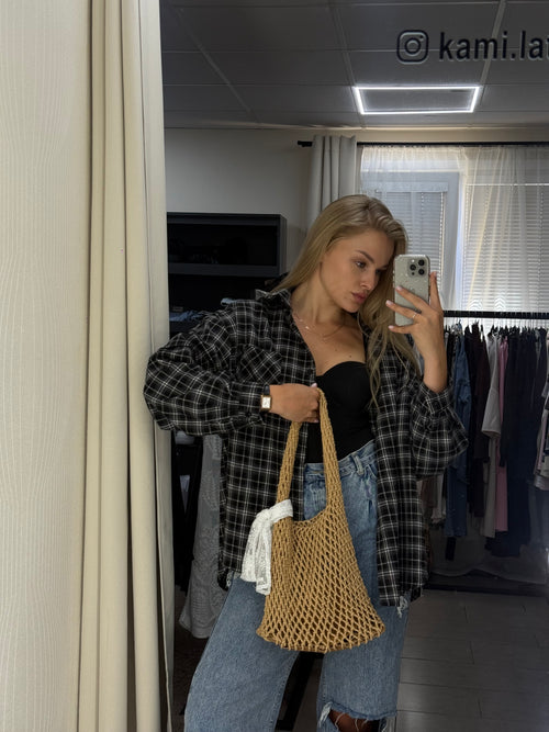 A woman takes a mirror selfie in a fitting room, wearing a black and white plaid flannel shirt, blue jeans, and holding a large woven straw shoulder bag.