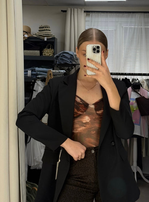 A woman takes a mirror selfie in a fitting room, wearing a black blazer over a brown and bronze patterned camisole top and dark brown pants.
