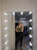 A person taking a mirror selfie in a fitting room, wearing a green sleeveless crop top and black leggings. The mirror is surrounded by bright light bulbs, and a clothing rack is visible in the background.