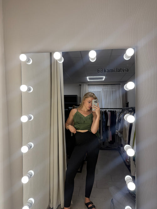 A person taking a mirror selfie in a fitting room, wearing a green sleeveless crop top and black leggings. The mirror is surrounded by bright light bulbs, and a clothing rack is visible in the background.