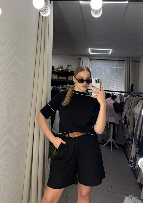 A woman takes a mirror selfie in a fitting room wearing a black short-sleeve mock-neck top with white piping and matching black shorts, accessorized with black sunglasses and a brown belt.