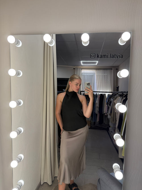 A woman takes a mirror selfie in a fitting room wearing a black sleeveless top and a beige maxi skirt. The mirror is surrounded by bright light bulbs, and clothing racks are visible in the background.
