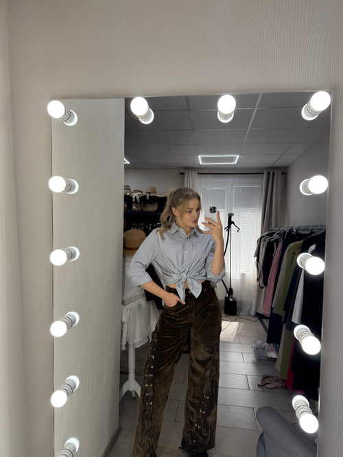 A person taking a mirror selfie in a dressing room, wearing a light gray long-sleeve button-up shirt and brown corduroy pants with silver stud embellishments. The mirror is surrounded by bright light bulbs, reflecting a closet with clothing racks and a white door in the background.