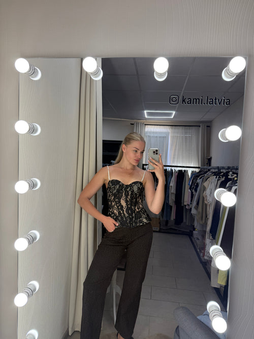 A woman takes a mirror selfie in a fitting room, wearing a black lace camisole top and dark brown pants. The image is upside down, showing a lighted vanity mirror with lights around the edge, a clothing rack, and a gray tiled wall in the background.