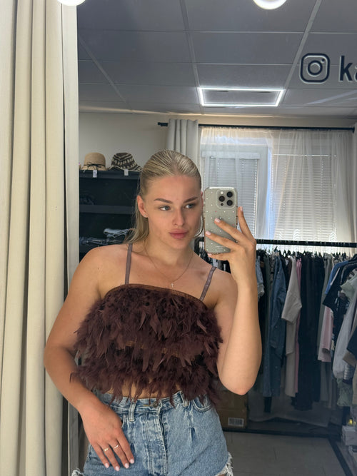 A woman taking a mirror selfie in a fitting room, wearing a dark brown feathered camisole top and denim shorts.