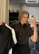 A woman takes a mirror selfie in a boutique, wearing a dark brown short-sleeve button-up shirt tied at the waist. She holds up a black shirt on a hanger in her left hand and a white shirt on a hanger in her right hand, with clothing racks and hats visible in the background.