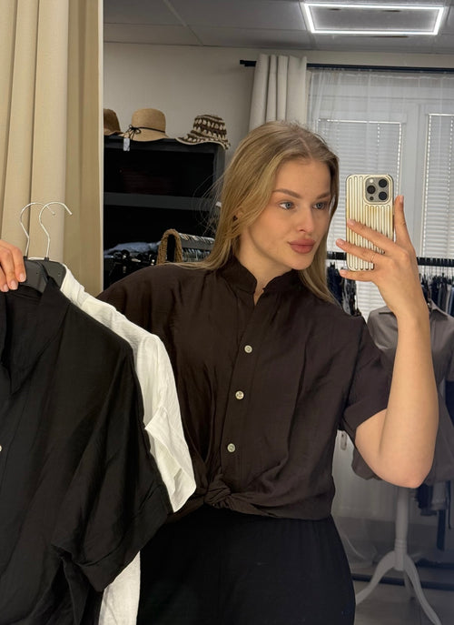 A woman takes a mirror selfie in a boutique, wearing a dark brown short-sleeve button-up shirt tied at the waist. She holds up a black shirt on a hanger in her left hand and a white shirt on a hanger in her right hand, with clothing racks and hats visible in the background.