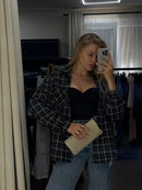 A woman takes a mirror selfie in a room, wearing a black and white plaid button-up shirt over a black top, blue jeans, and holding a glittery gold clutch.