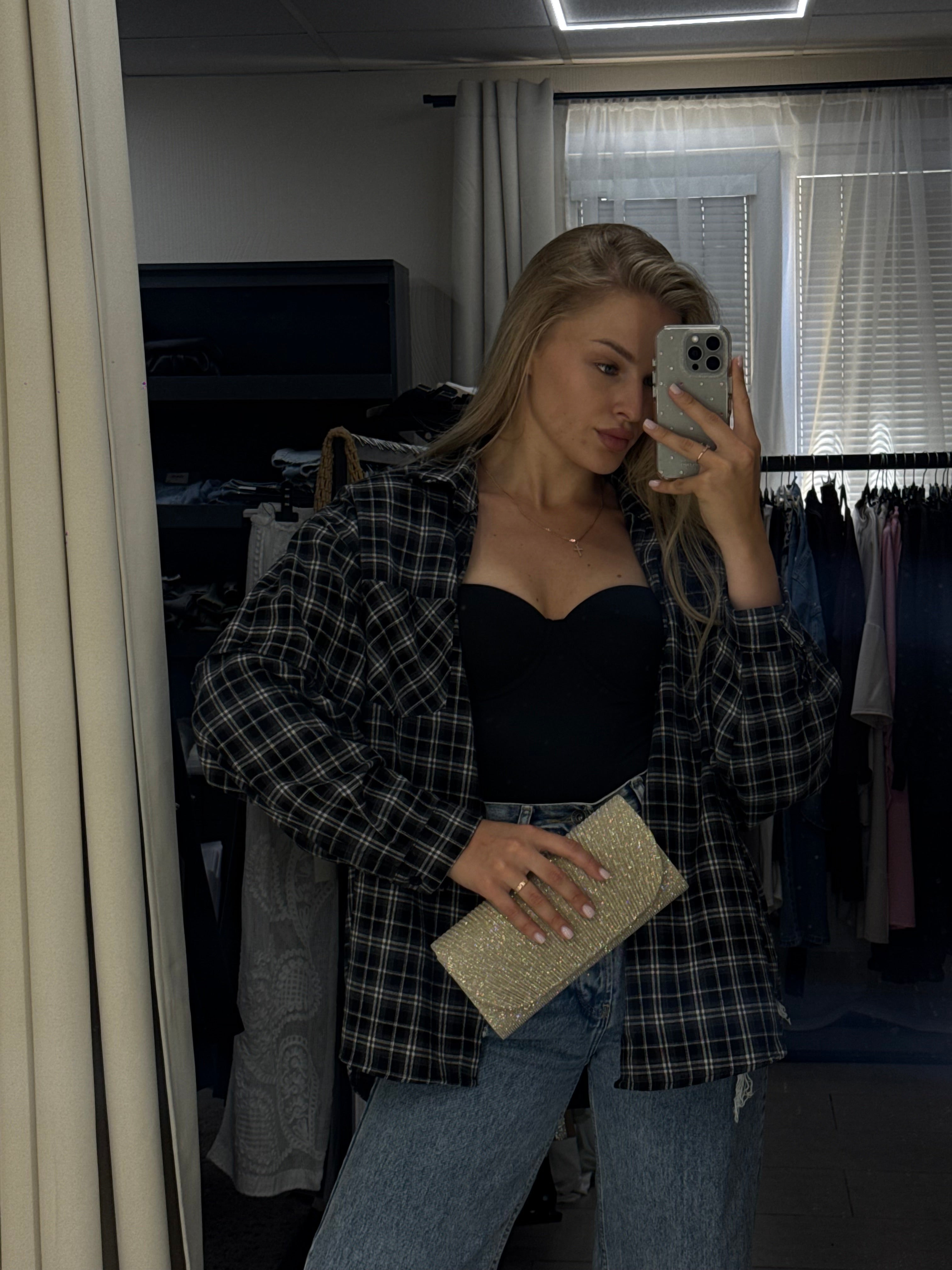 A woman takes a mirror selfie in a room, wearing a black and white plaid button-up shirt over a black top, blue jeans, and holding a glittery gold clutch.