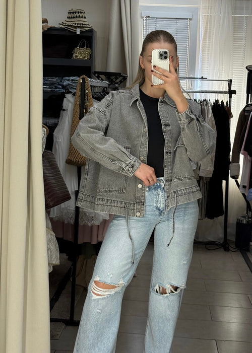 A person taking a mirror selfie in a clothing boutique, wearing a faded gray denim jacket over a black top and light-wash distressed jeans.