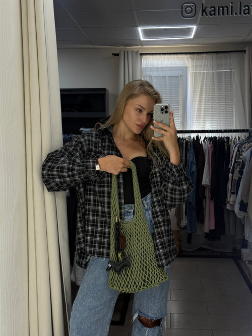 A woman takes a mirror selfie in a fitting room or closet area, wearing a black and white plaid flannel shirt, light blue jeans, and carrying a green mesh tote bag.