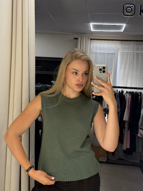 A woman taking a mirror selfie in a room, wearing a sleeveless ribbed knit top in sage green. The background shows a closet with clothes hanging on a rack and a lighted mirror frame.