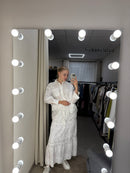 A woman takes a mirror selfie wearing a white long-sleeve lace dress in what appears to be a dressing room or closet, with a lighted vanity mirror showing her reflection.