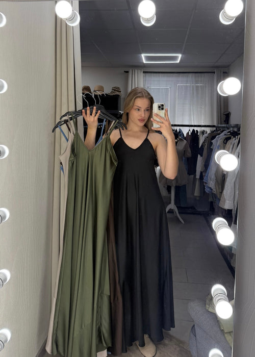 A woman takes a mirror selfie in a dressing room, wearing a long black spaghetti-strap maxi dress. She holds up two other dresses on hangers: an olive green one and a dark brown one.