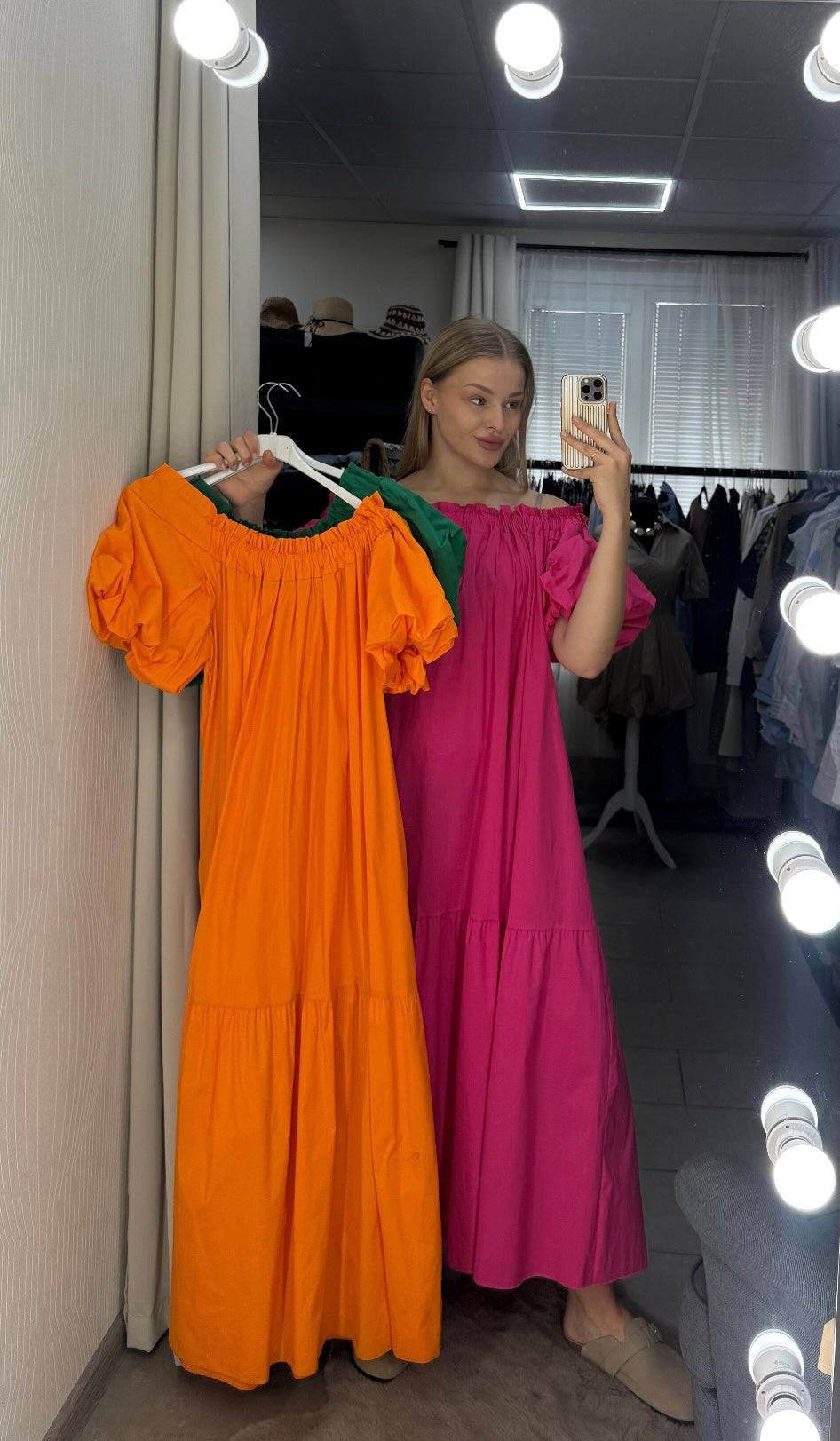 Woman taking a mirror selfie wearing a pink dress next to an orange dress on a hanger.