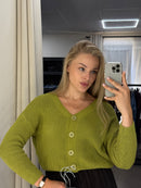 A woman takes a mirror selfie wearing a chunky olive green knit cardigan sweater with cream buttons. The photo is taken in a room with a mirror, showing a window and light fixture in the background.