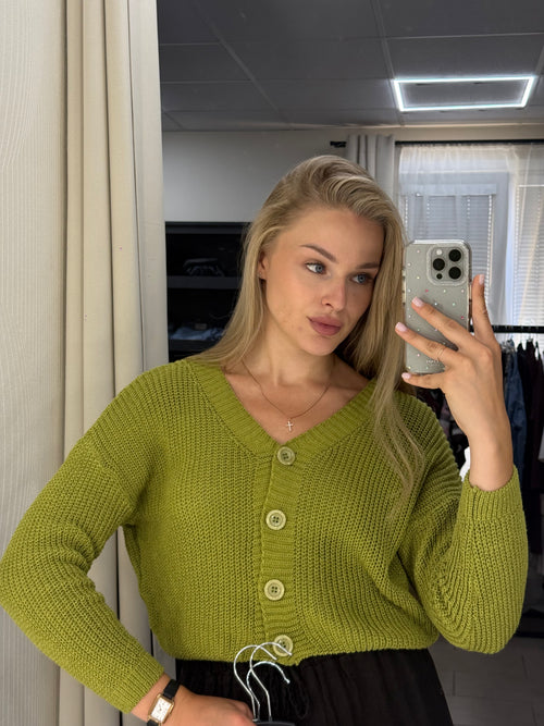 A woman takes a mirror selfie wearing a chunky olive green knit cardigan sweater with cream buttons. The photo is taken in a room with a mirror, showing a window and light fixture in the background.