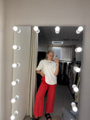 A person taking a mirror selfie in a fitting room, wearing a white t-shirt and bright red wide-leg pants. The image is rotated 90 degrees counter-clockwise, showing a lighted vanity mirror with bulbs around the edge.