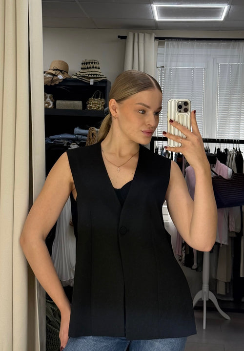 A woman taking a mirror selfie in a fitting room wearing a sleeveless black vest over a black top with blue jeans.