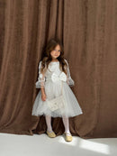 Young girl in a white dress with a large bow standing against a brown curtain.