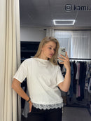 A woman takes a mirror selfie in a fitting room wearing a white short-sleeve top with a lace hem and black pants. The room has a mirror, clothing rack, and window blinds in the background.