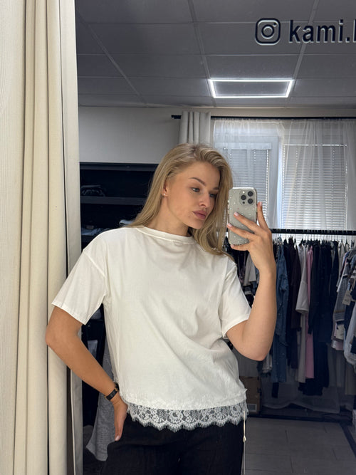 A woman takes a mirror selfie in a fitting room wearing a white short-sleeve top with a lace hem and black pants. The room has a mirror, clothing rack, and window blinds in the background.