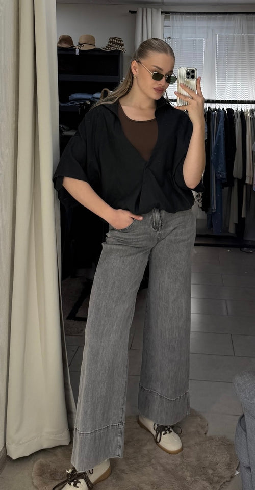 A woman takes a mirror selfie in a fitting room or closet area, wearing a black short-sleeve wrap-style top over a brown crew-neck shirt, paired with wide-leg gray jeans and white sneakers.