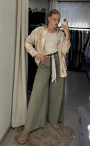 A woman takes a mirror selfie in a fitting room or closet area, wearing a light beige satin bomber jacket, a cream ribbed tank top, wide-leg olive green pants, and beige slip-on shoes.