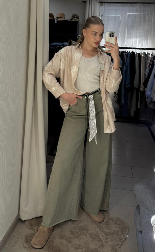 A woman takes a mirror selfie in a fitting room or closet area, wearing a light beige satin bomber jacket, a cream ribbed tank top, wide-leg olive green pants, and beige slip-on shoes.