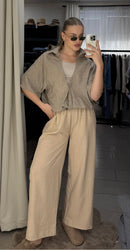 A woman takes a mirror selfie in a fitting room or closet area, wearing a loose taupe short-sleeve button-up shirt over a white tank top and beige wide-leg trousers. She is wearing sunglasses and slippers.